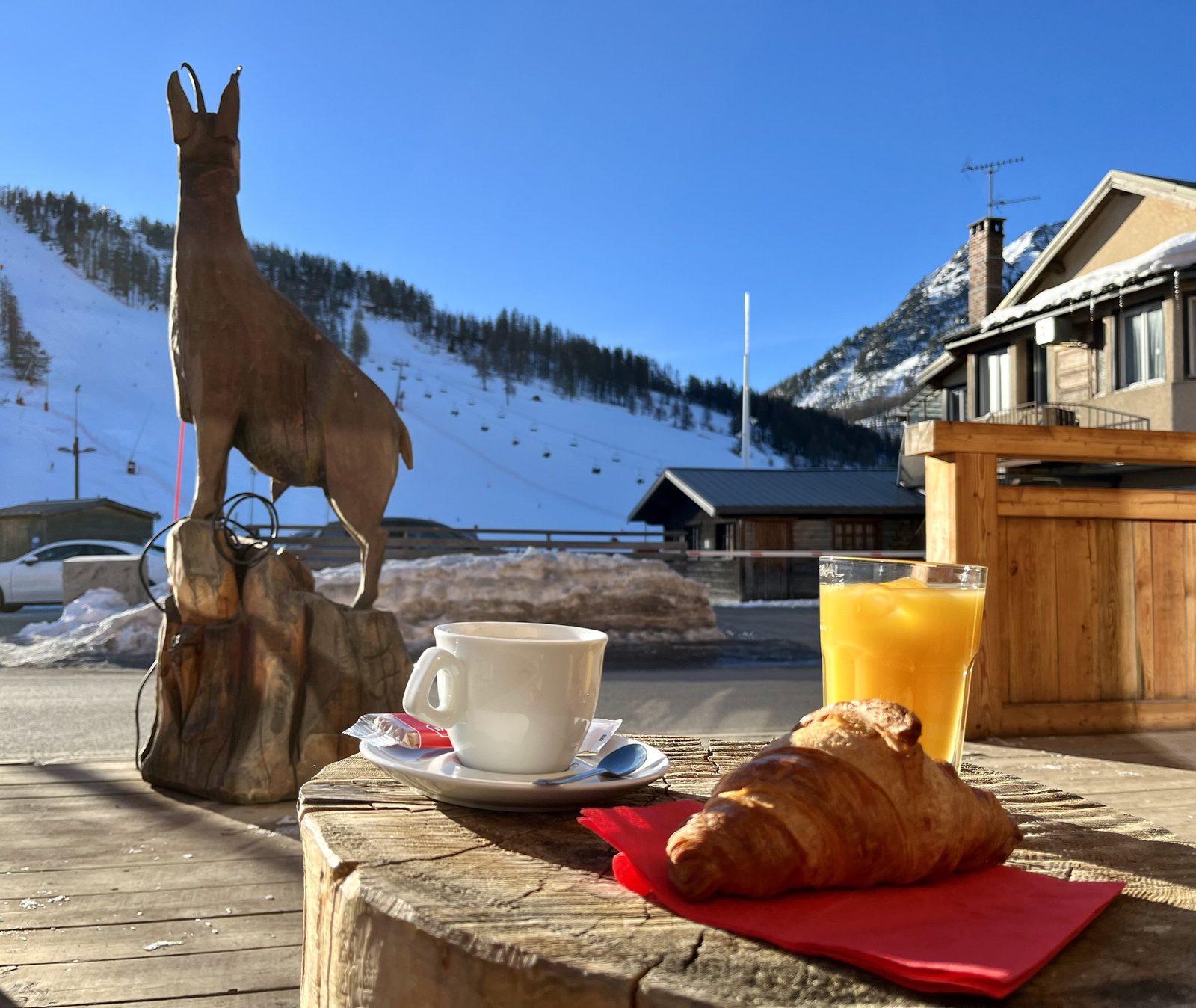 Un café, un croissant et un jus d’orange sur une table en bois, une piste de ski alpine en arrière-plan.